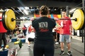 Woman standing in a gym holding a heavily loaded barbell at shoulder height in preparation for an overhead weightlifting movement
