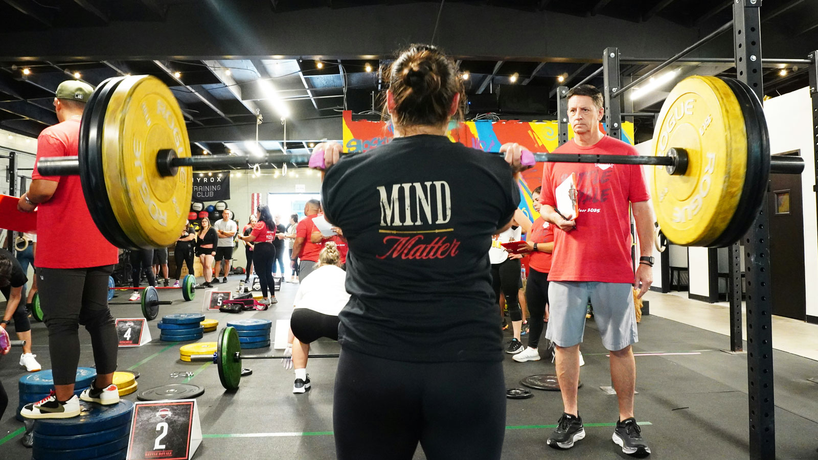 Woman standing in a gym holding a heavily loaded barbell at shoulder height in preparation for an overhead weightlifting movement