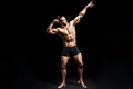 Muscular male bodybuilder wearing posing briefs with one arm extended upward and the other bent to flex his biceps while looking toward his raised hand