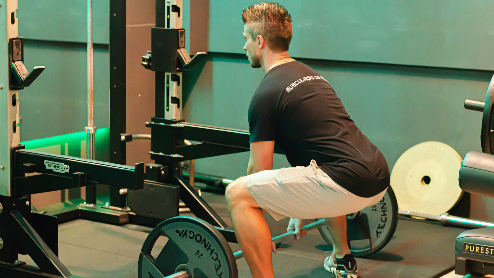 Man positioned in a squat near a gym bench preparing to lift weights while maintaining a controlled exercise posture