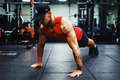 Man wearing a red tank top and black shorts performing push-ups on the gym floor during a strength-training workout