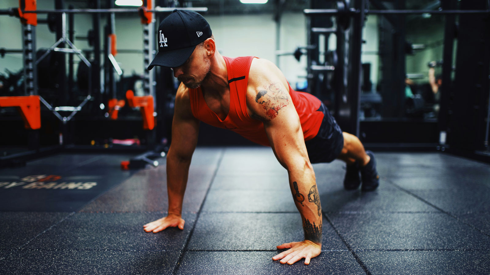 Man wearing a red tank top and black shorts performing push-ups on the gym floor during a strength-training workout