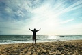 Person standing on a beach at sunset with both arms raised toward the sky in a gesture of celebration and personal achievement