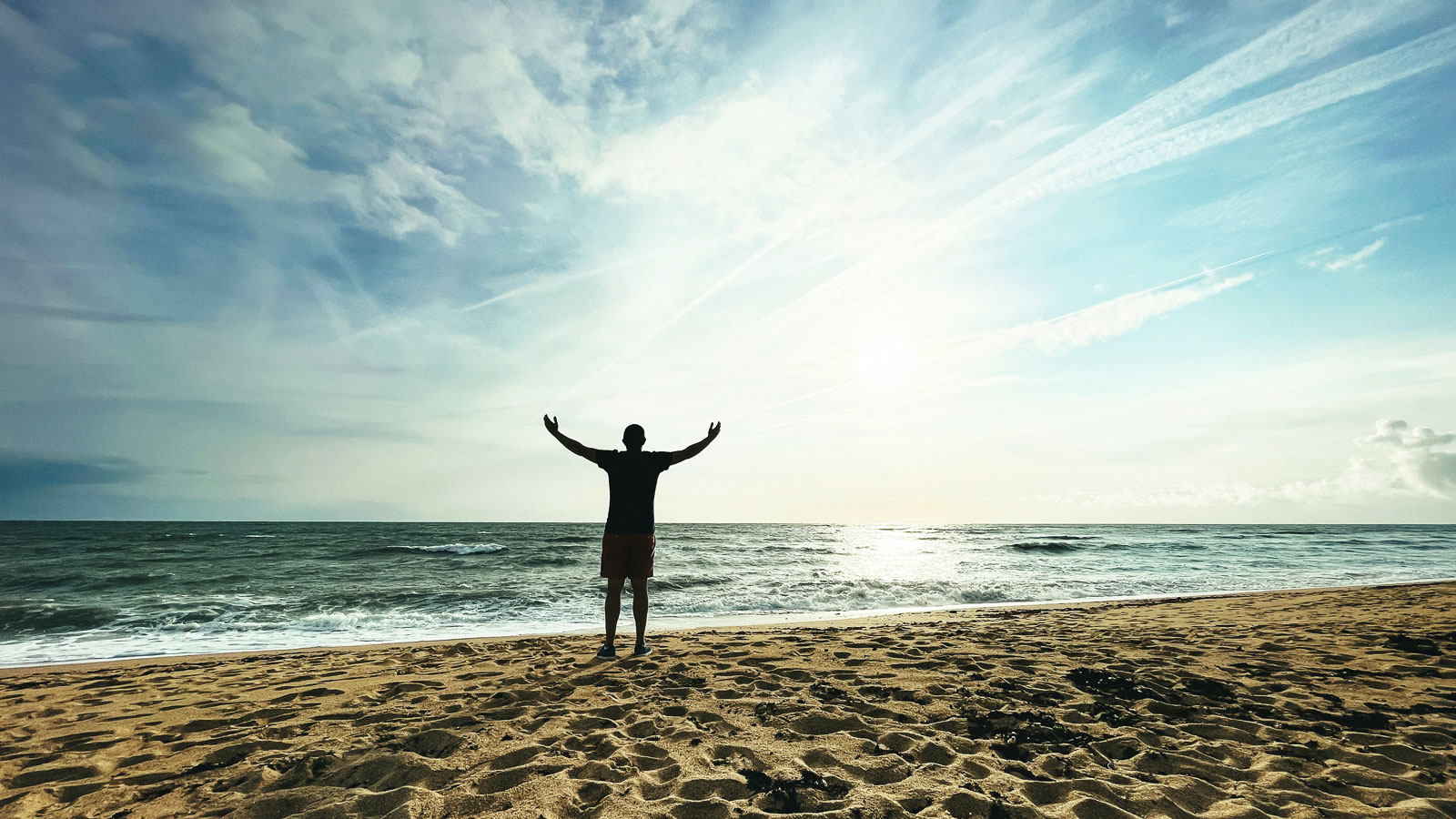 Person standing on a beach at sunset with both arms raised toward the sky in a gesture of celebration and personal achievement