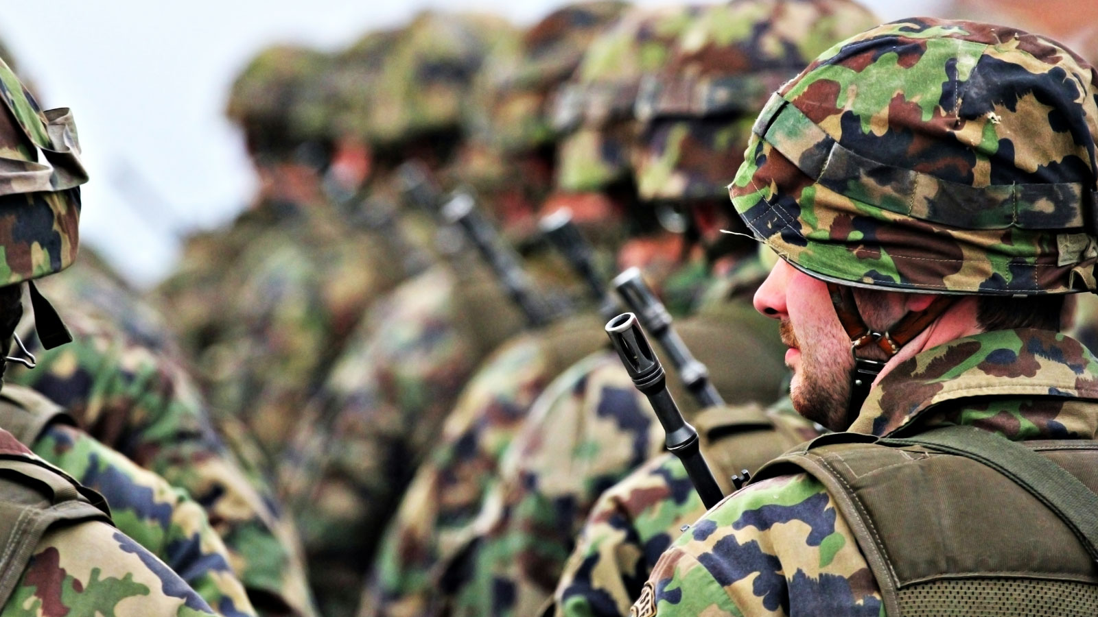 Side view of Swiss soldiers standing in formation with a foreground soldier wearing a camouflage helmet and tactical vest while holding a rifle pointed upward